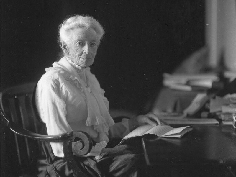 ellen browning scripps in white blouse and dark skirt sitting in chair at her desk looking at camera