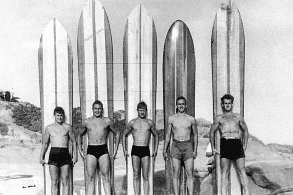 Pioneer surfers in 1946 stand on Windansea beach with their long surfboards behind them, standing up in the sand