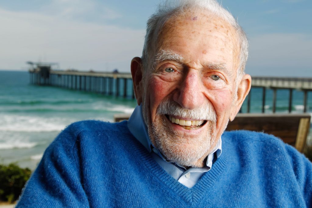 photo of the late oceanographer Walter munk, he wears a blue sweater and the scripps pier is behind him