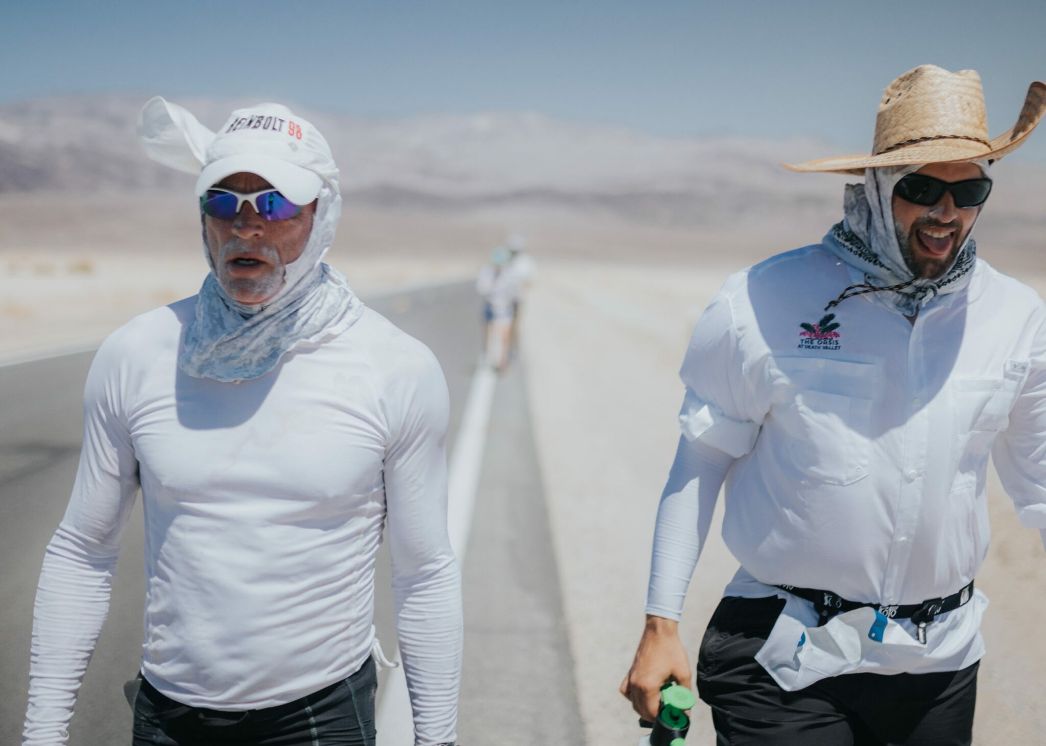 two men in white shirts and sun hats walk on the side of the street in Death Valley