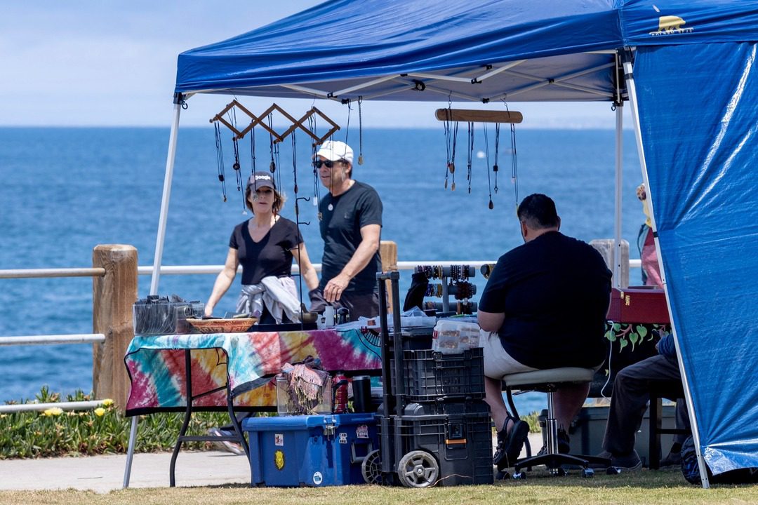 visitors walk by sidewalk vending in La Jolla where a table is set up with merchandise for sale and blue pop up tent over the table
