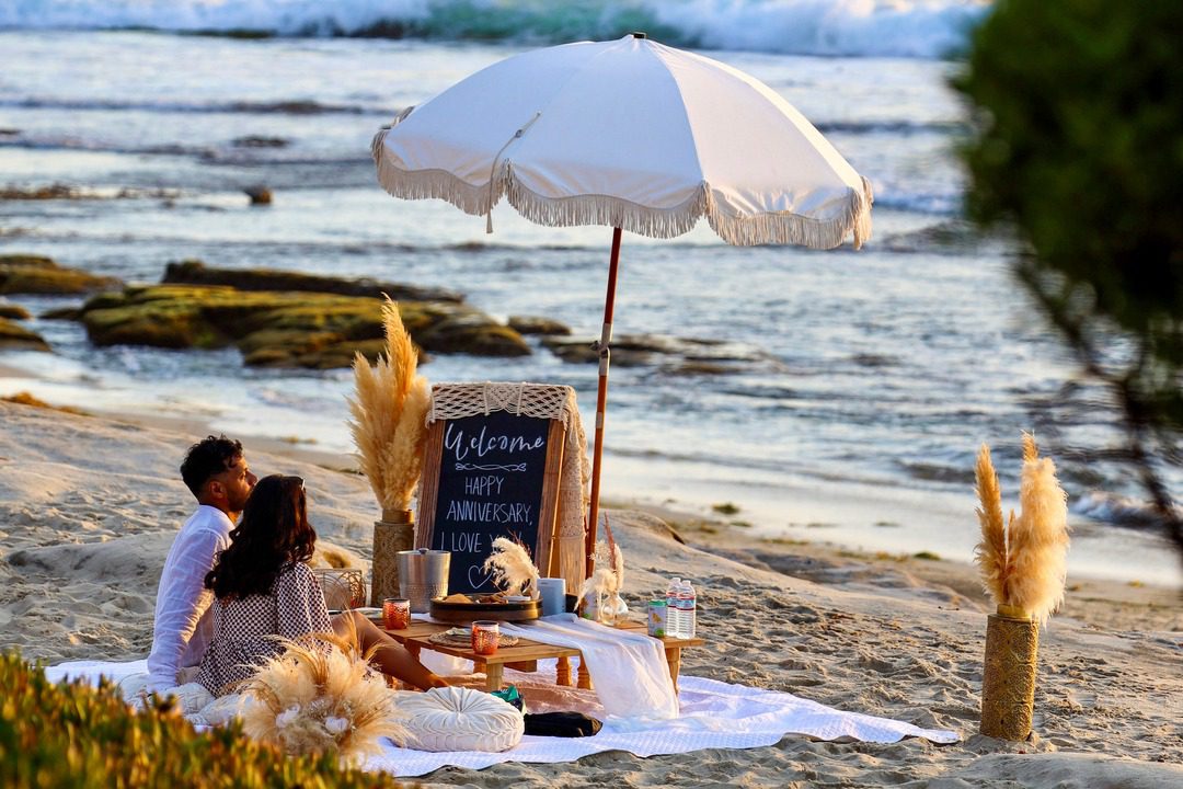 a man and woman sit on the beach looking out at the ocean. the luxury picnic is set up around them with an umbrella, a happy anniversary sign, food and drink, and other decor