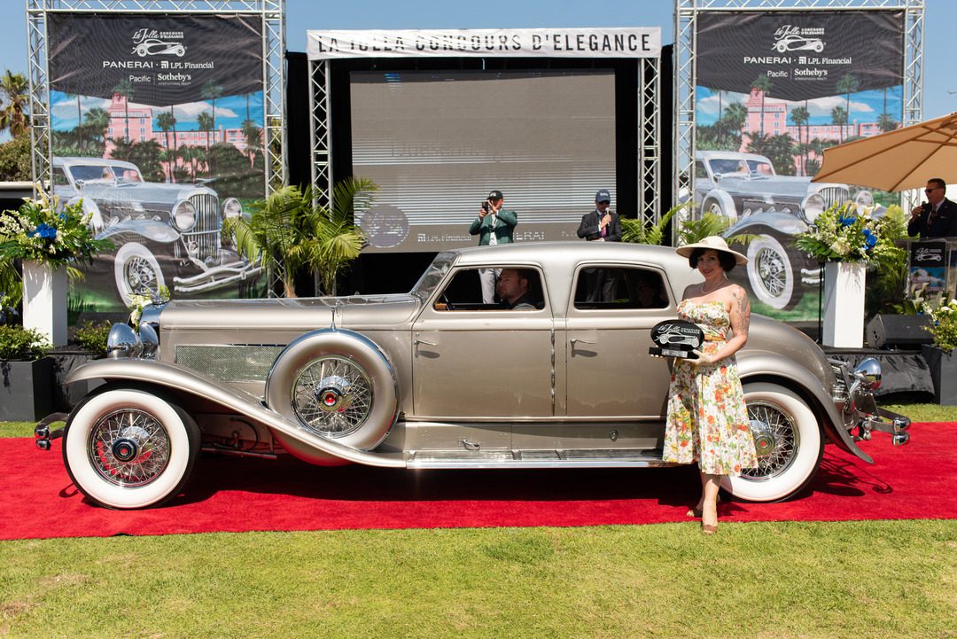 woman in dress and hat holds trophy in front of a classic car at the La Jolla concours d'elegance showcase