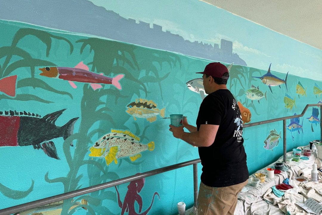 artist amadeo bachar stands in front of mural painting underwater scene at the children's school