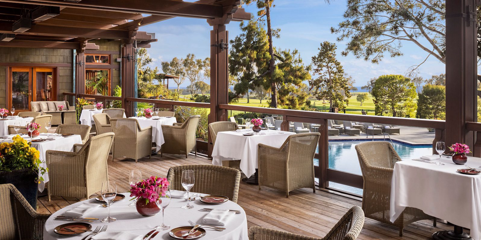 A beautifully set outdoor terrace dining area at the A.R. Valentien restaurant, featuring wicker chairs and round tables covered with white tablecloths. Each table is adorned with vibrant flower arrangements and neatly arranged silverware and glassware. The terrace overlooks a serene landscape with lush greenery, a glimpse of a golf course, and a swimming pool in the background. The wooden beams and rustic design elements of the terrace enhance the charming and relaxed atmosphere, offering guests an inviting setting to enjoy a meal with a view of the distant trees and ocean