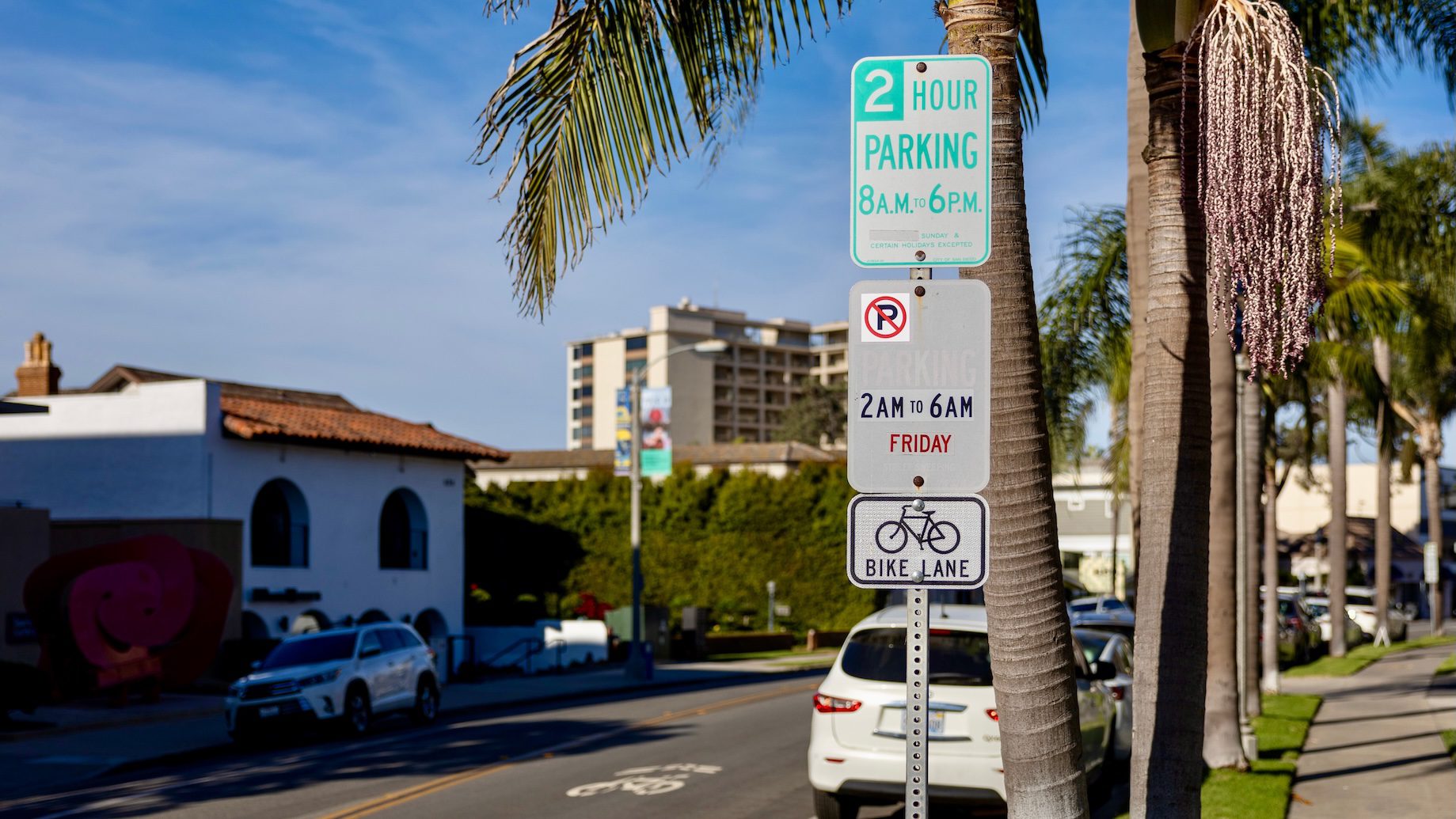 Street view of La Jolla parking signs indicating 2-hour parking from 8 AM to 6 PM, no parking from 2 AM to 6 AM on Fridays, and a bike lane, with palm trees and buildings in the background.