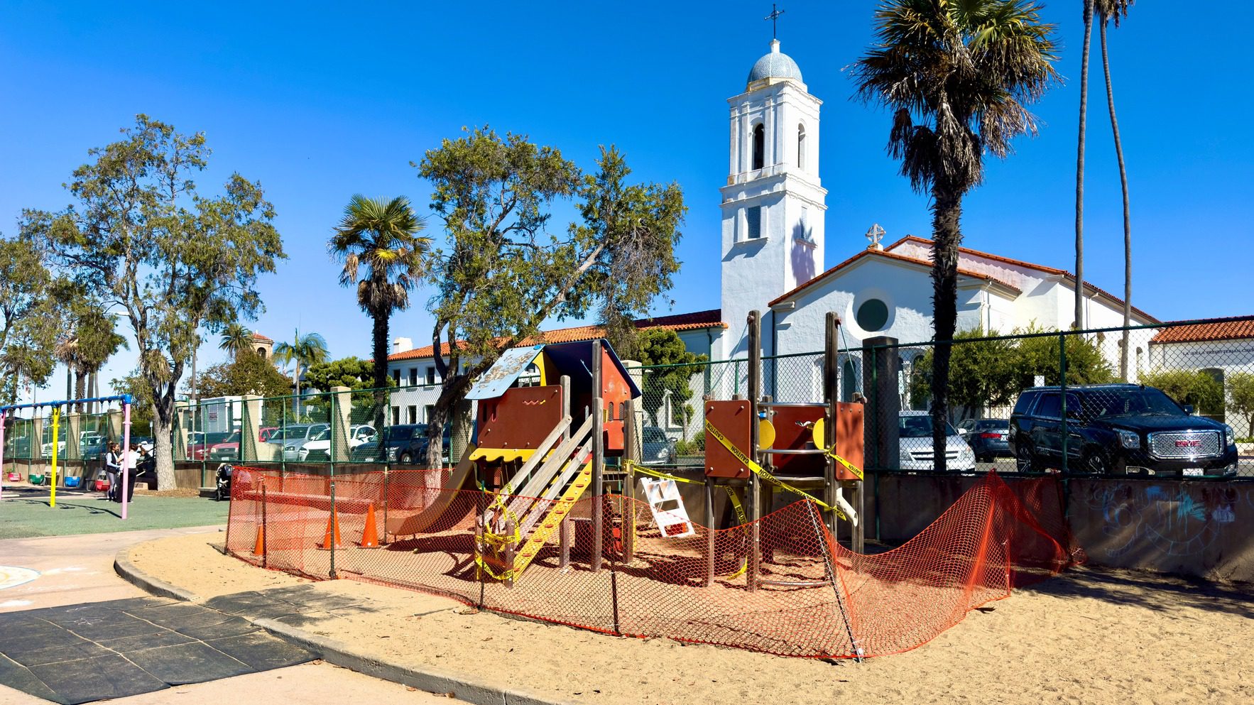 Rec center playground in La Jolla surrounded by caution tape, featuring play structures and a fenced area, with a church and parked cars in the background on a clear, sunny day.
