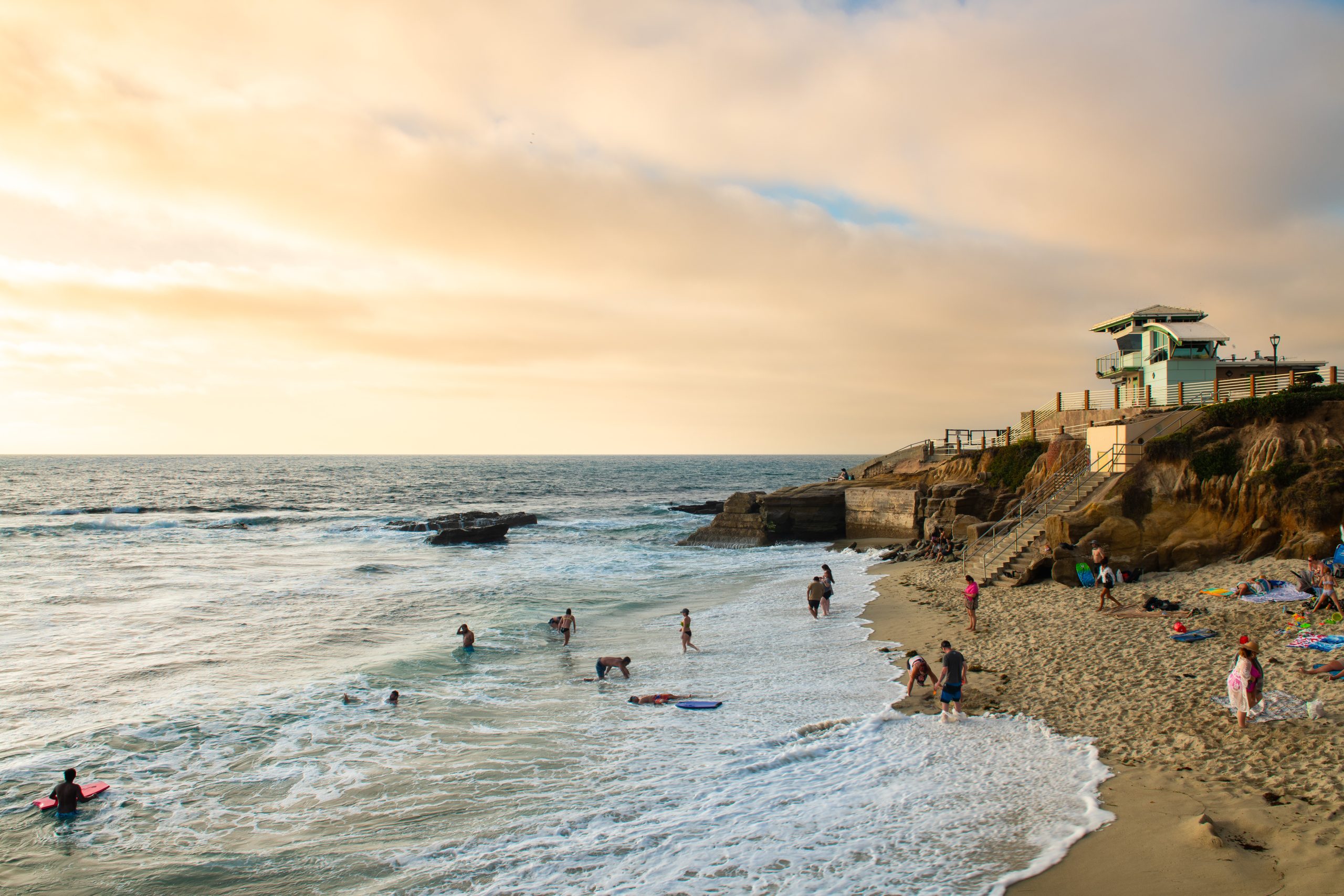 Sunset view of South Casa Beach in La Jolla, CA, with families playing in the surf, sandstone cliffs, and a lifeguard tower above the shoreline.