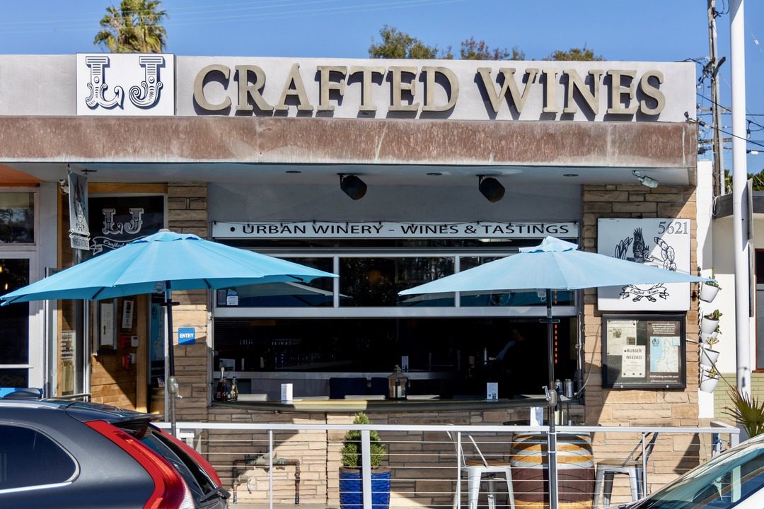 Front view of LJ Crafted Wines in Bird Rock, San Diego, featuring a rustic storefront, blue patio umbrellas, and signage for wine tastings at this urban winery.
