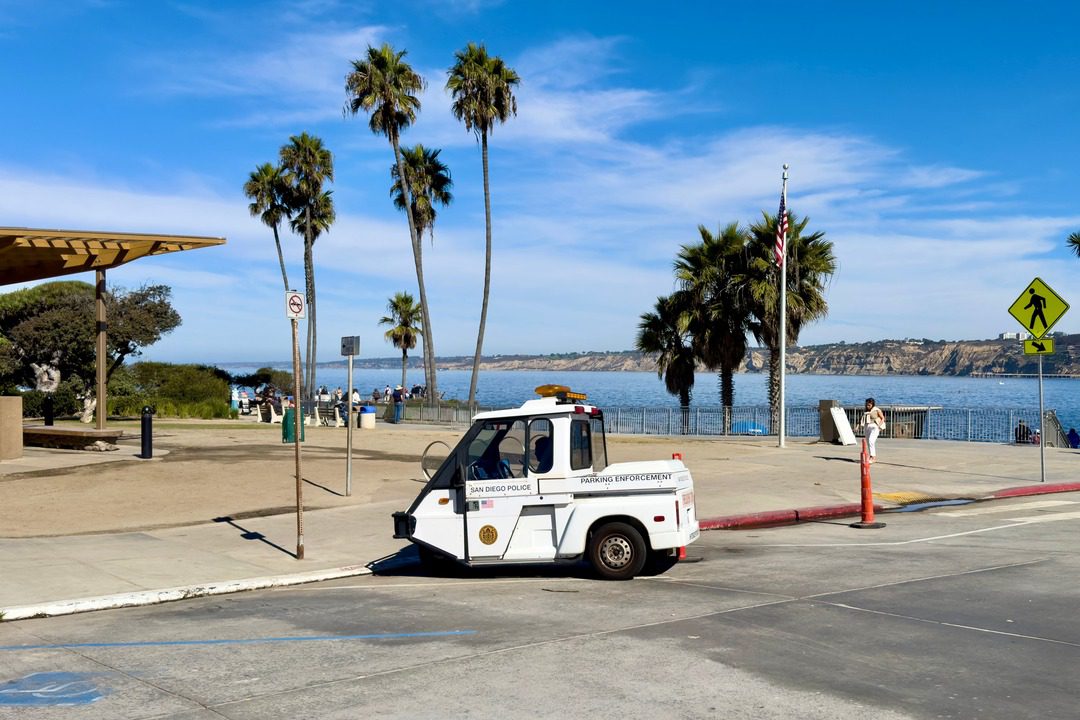San Diego Police parking enforcement vehicle parked near La Jolla Cove with a scenic ocean view, palm trees, an American flag, and a pedestrian crossing sign in the background.