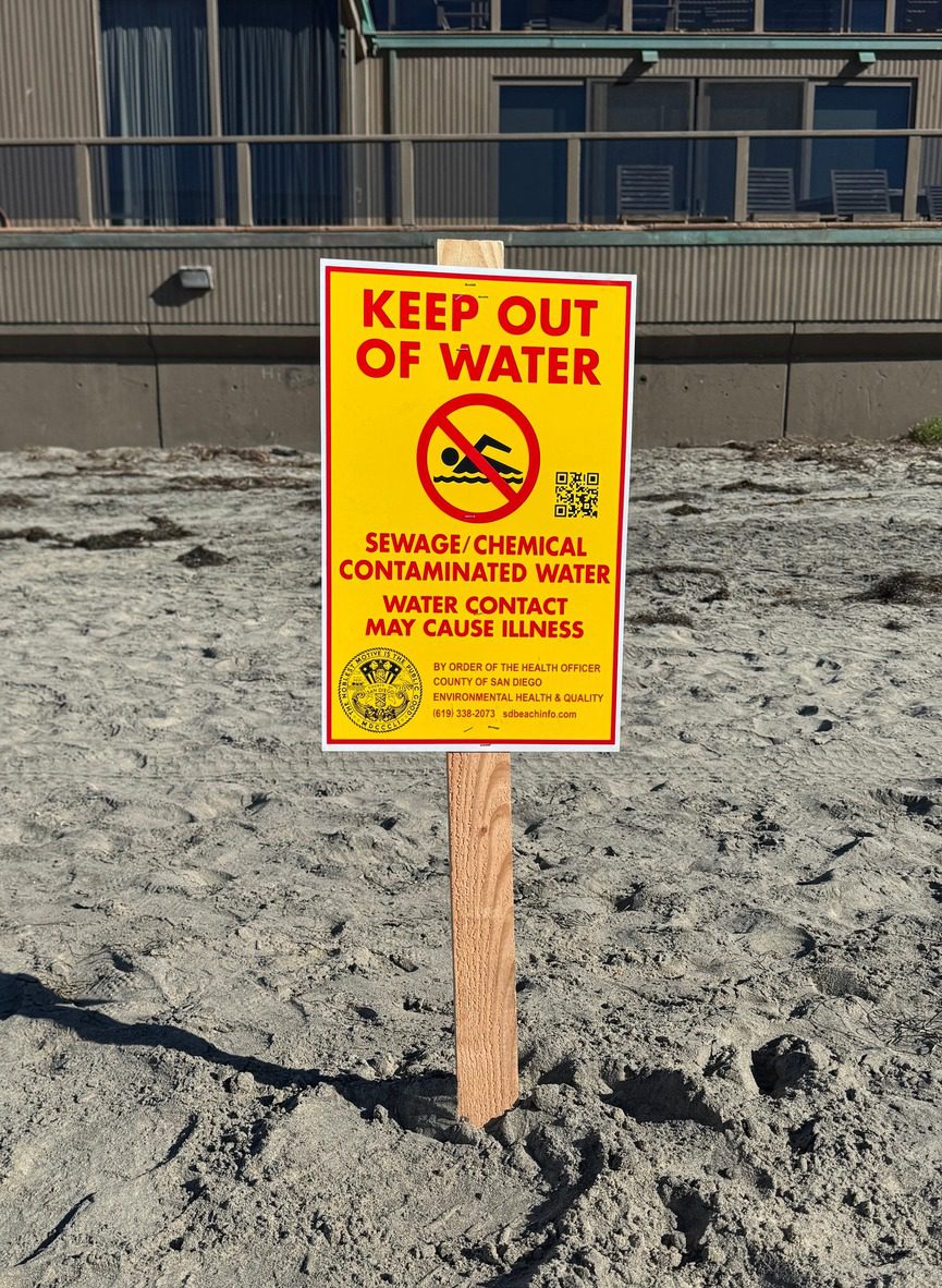 Close-up of a warning sign at La Jolla Shores Beach advising against water contact due to sewage and chemical contamination from a spill.