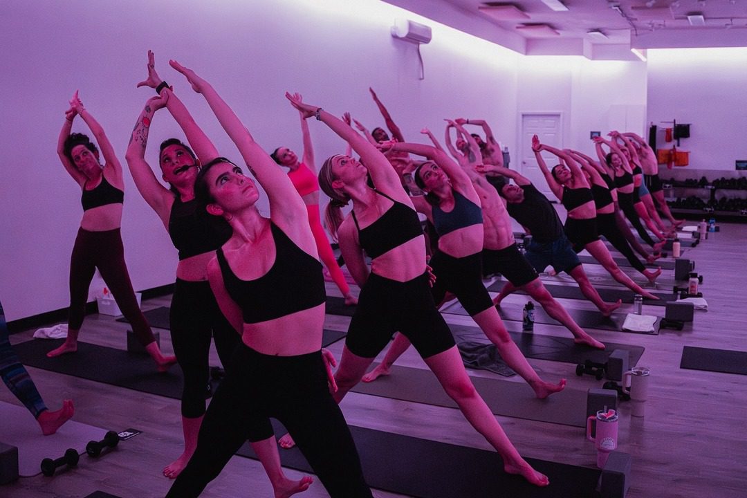 Group of individuals practicing yoga in a serene, purple-lit studio at Yoga Box La Jolla. Perfectly capturing the essence of "yoga in La Jolla," the participants are in a side stretch pose, showcasing focus and balance in a supportive, vibrant atmosphere.