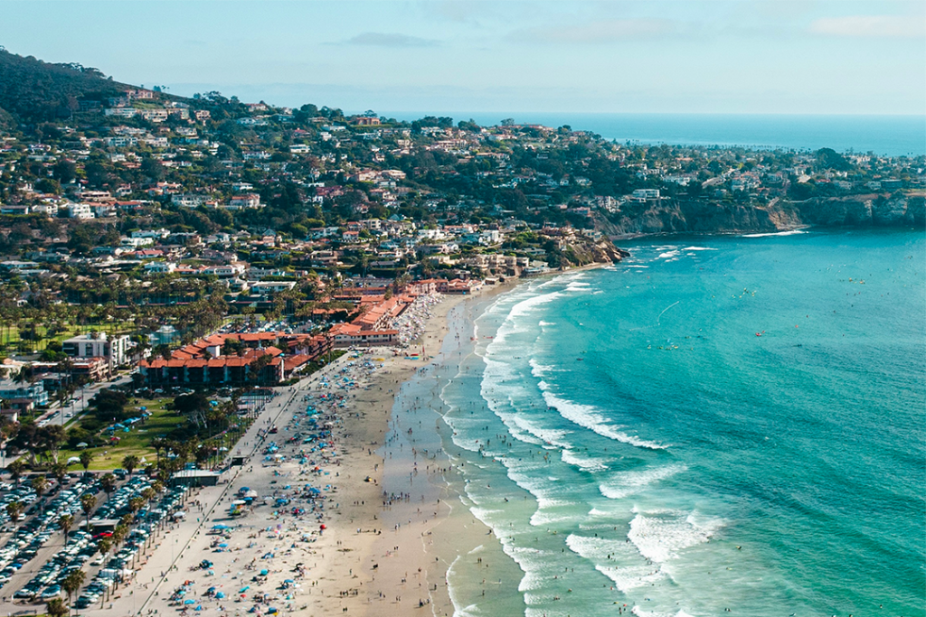 Aerial view of La Jolla Shores Beach in La Jolla, CA, with crowds along the shoreline, turquoise waves, and hillside homes in the background.