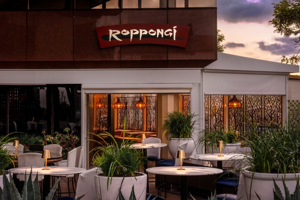 Outdoor patio seating at Roppongi restaurant La Jolla, featuring white tables, gold candle lamps, and lush greenery at dusk.