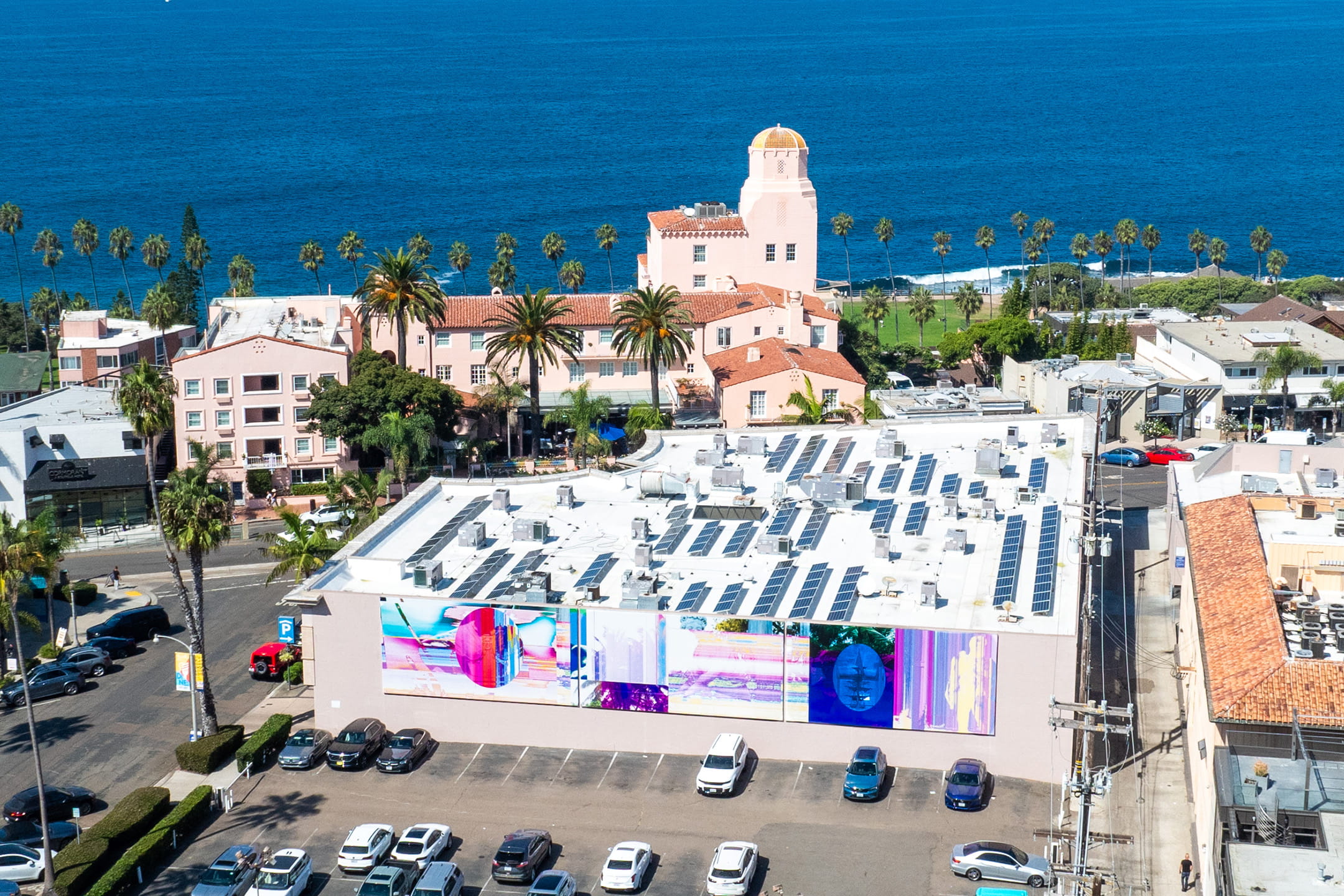 An aerial view of "Ghosts in the Machine" (2024) by Todd Gray, a vibrant triptych mural in iridescent pinks, blues, and greens on the back of a building at 1111 Prospect Street — part of the Murals of La Jolla public art program, with the iconic La Valencia Hotel and Pacific Ocean visible in the background.