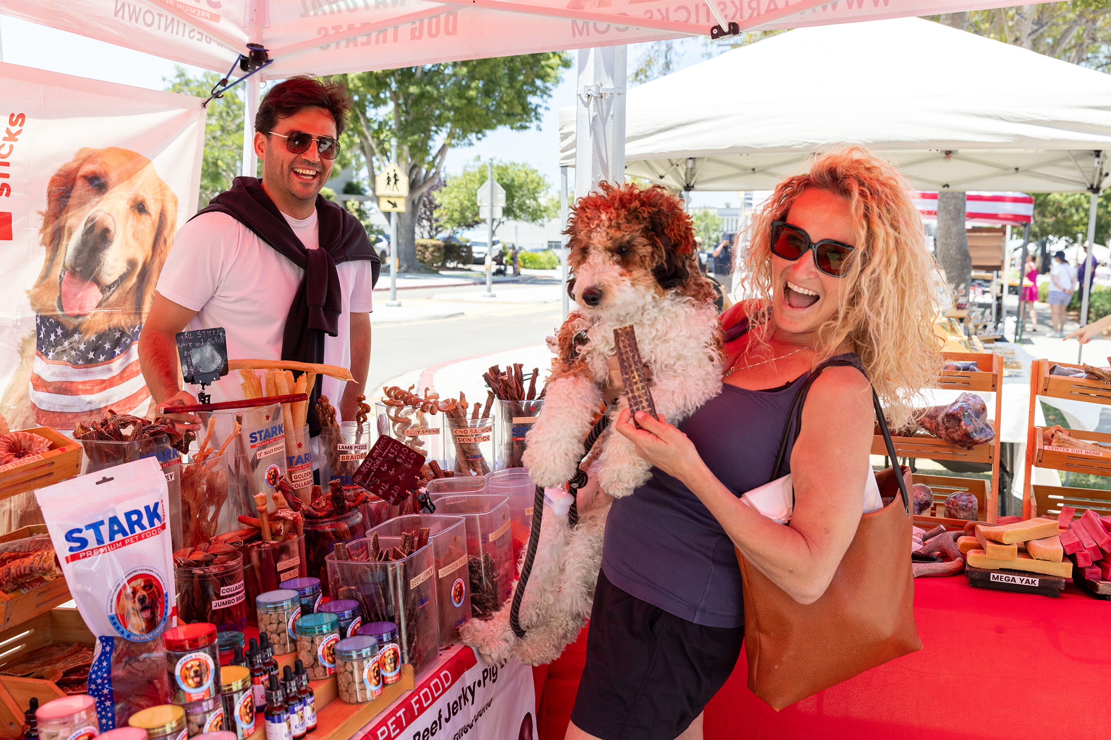 A woman holds her curly-haired dog while shopping for pet treats at the Stark Premium Pet Food booth at the La Jolla Open Aire Market.