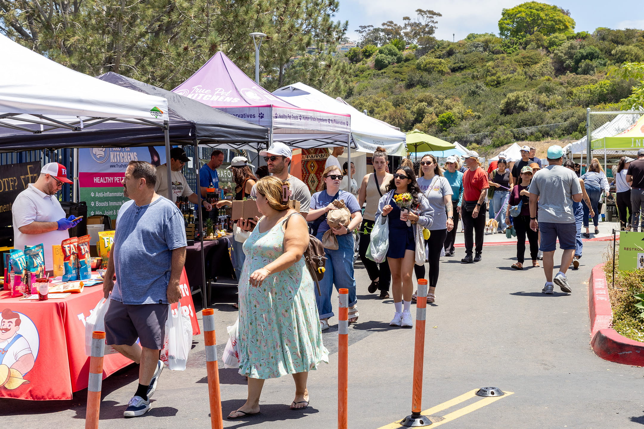 A crowd of shoppers strolls past rows of vendor tents at the La Jolla Open Aire Market on a sunny day with hillside views in the background.