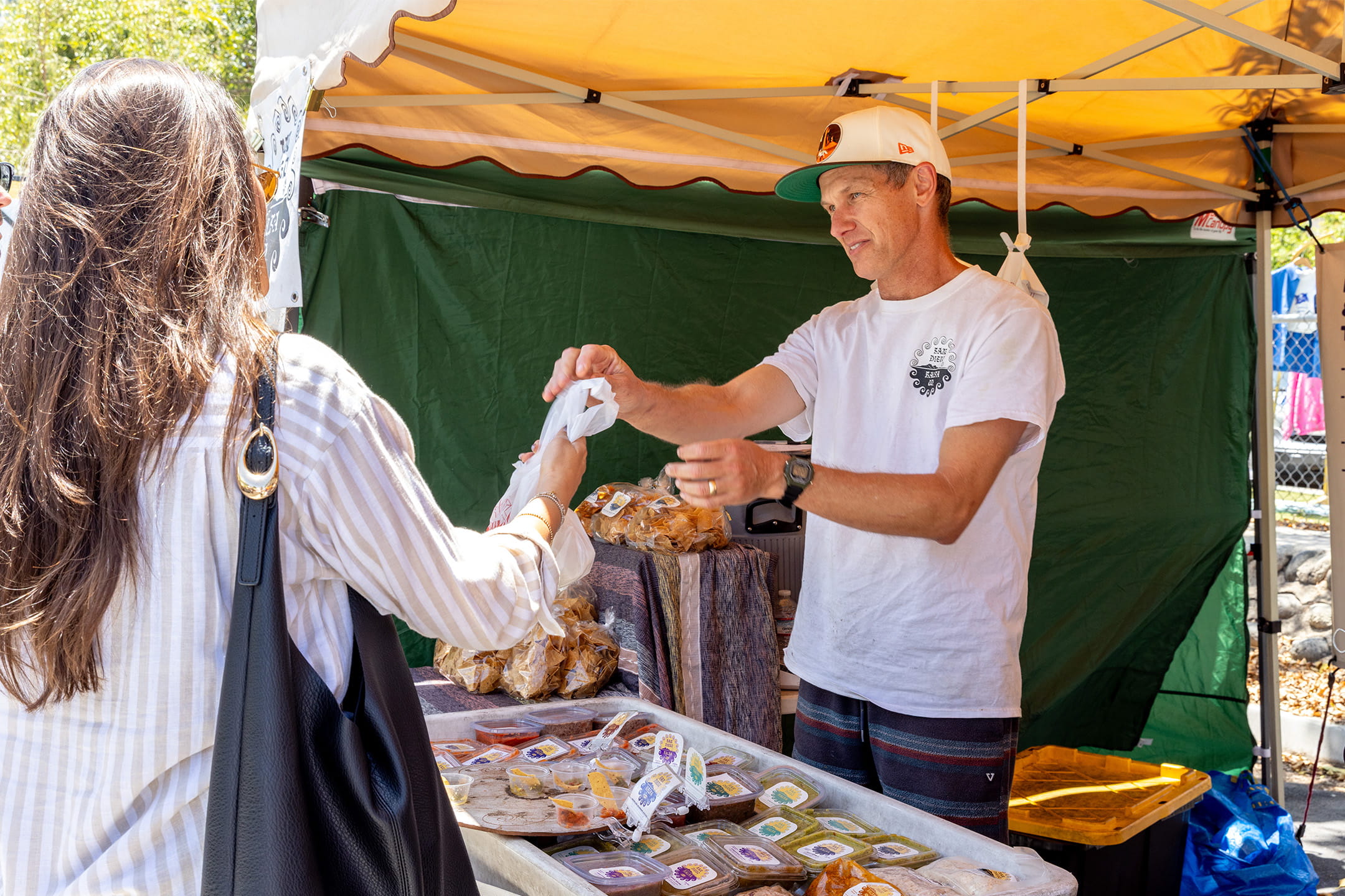 A vendor hands a bag of goods to a customer at a salsa and specialty food stand at the La Jolla Open Aire Market.
