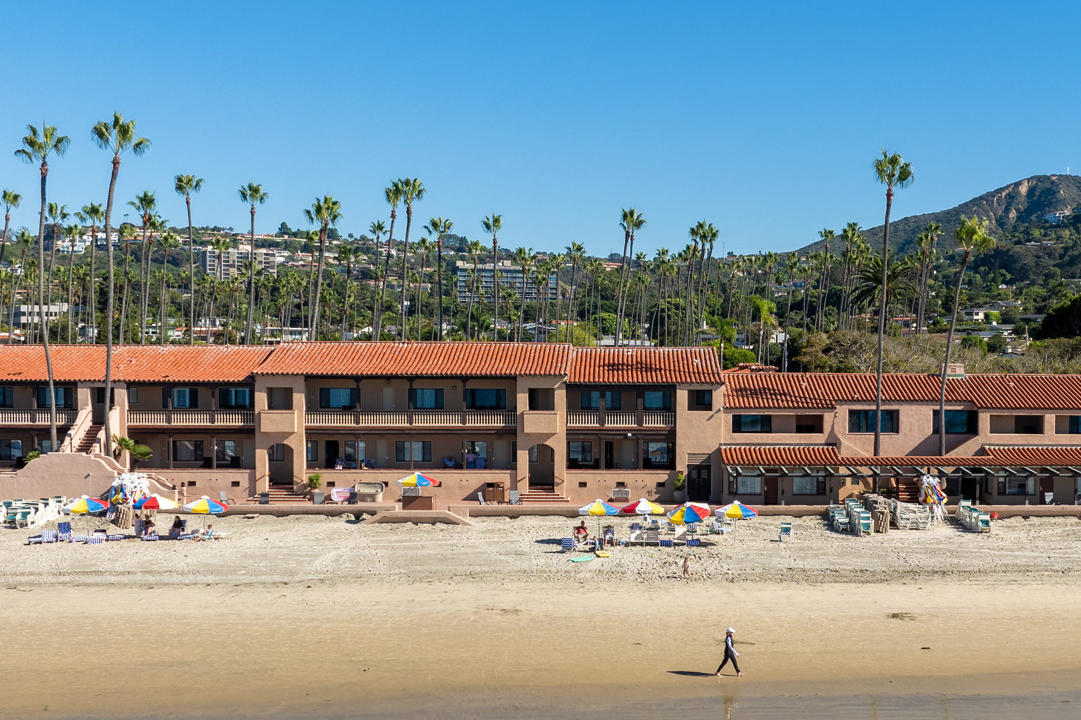 La Jolla luxury hotels beachfront villas with red tile roofs and palm trees, striped umbrellas and lounge chairs on the sand at low tide.