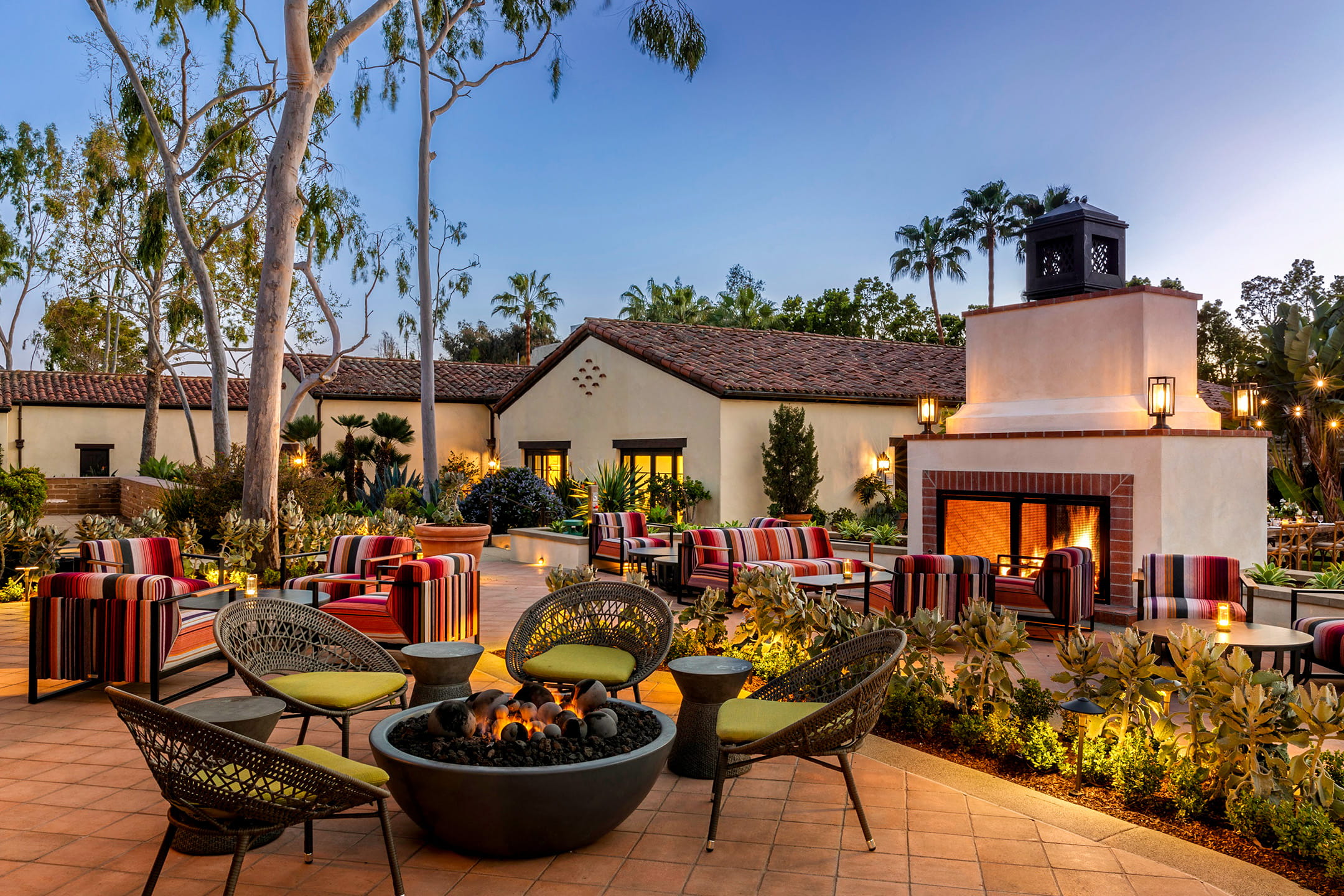 La Jolla luxury hotels outdoor lounge at twilight, striped patio chairs and a fire pit facing a lit fireplace beside a tiled Spanish-style building.