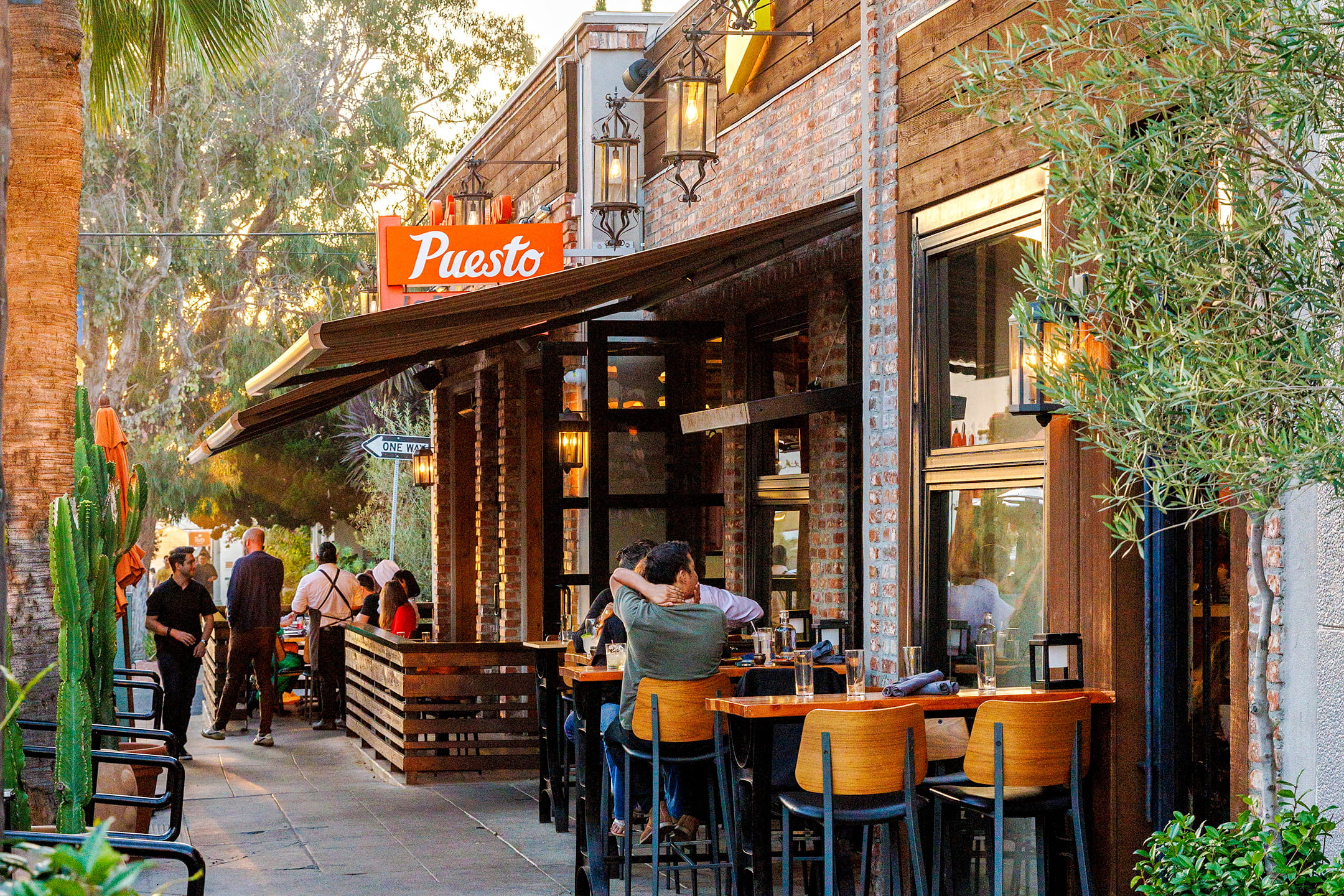 Outdoor dining in La Jolla at Puesto's Wall Street patio, with wooden bar seating and diners under the restaurant's brick facade and awning.